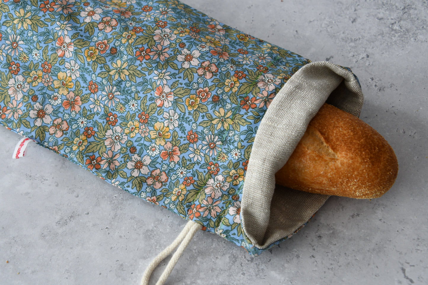 Ein handgenähter, langer Baguettebeutel liegt auf einer hellen Küchenarbeitsplatte. Ein frisches, knuspriges Baguette schaut oben leicht aus der geöffneten Kordelöffnung heraus.