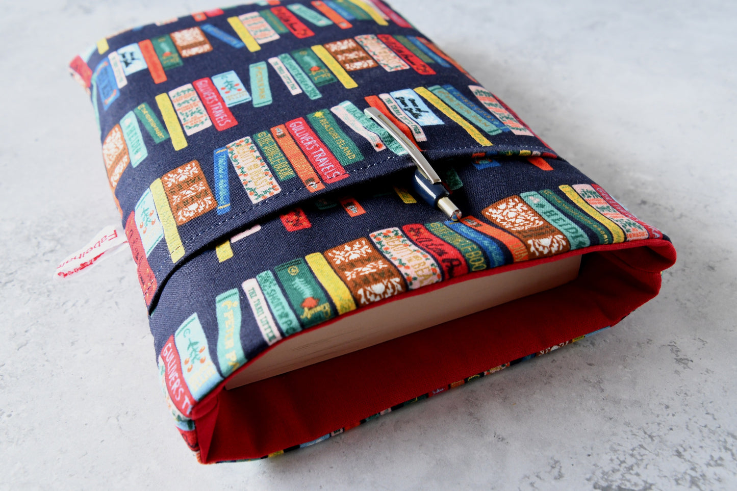 Detailreiches Foto einer Buchtasche mit Bücherrücken-Print. Aus der weichen Hülle schaut ein echtes Buch heraus. Das clevere Zubehörfach auf der Vorderseite ist mit einem Kugelschreiber bestückt, um den perfekten Schutz für unterwegs zu demonstrieren.
