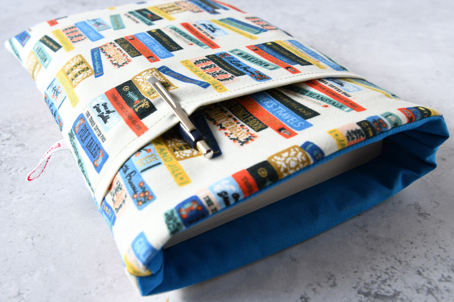 Detailreiches Foto einer Buchtasche mit Bücherrücken-Print. Aus der weichen Hülle schaut ein echtes Buch heraus. Das clevere Zubehörfach auf der Vorderseite ist mit einem Stift bestückt, um den perfekten Schutz für unterwegs zu demonstrieren.