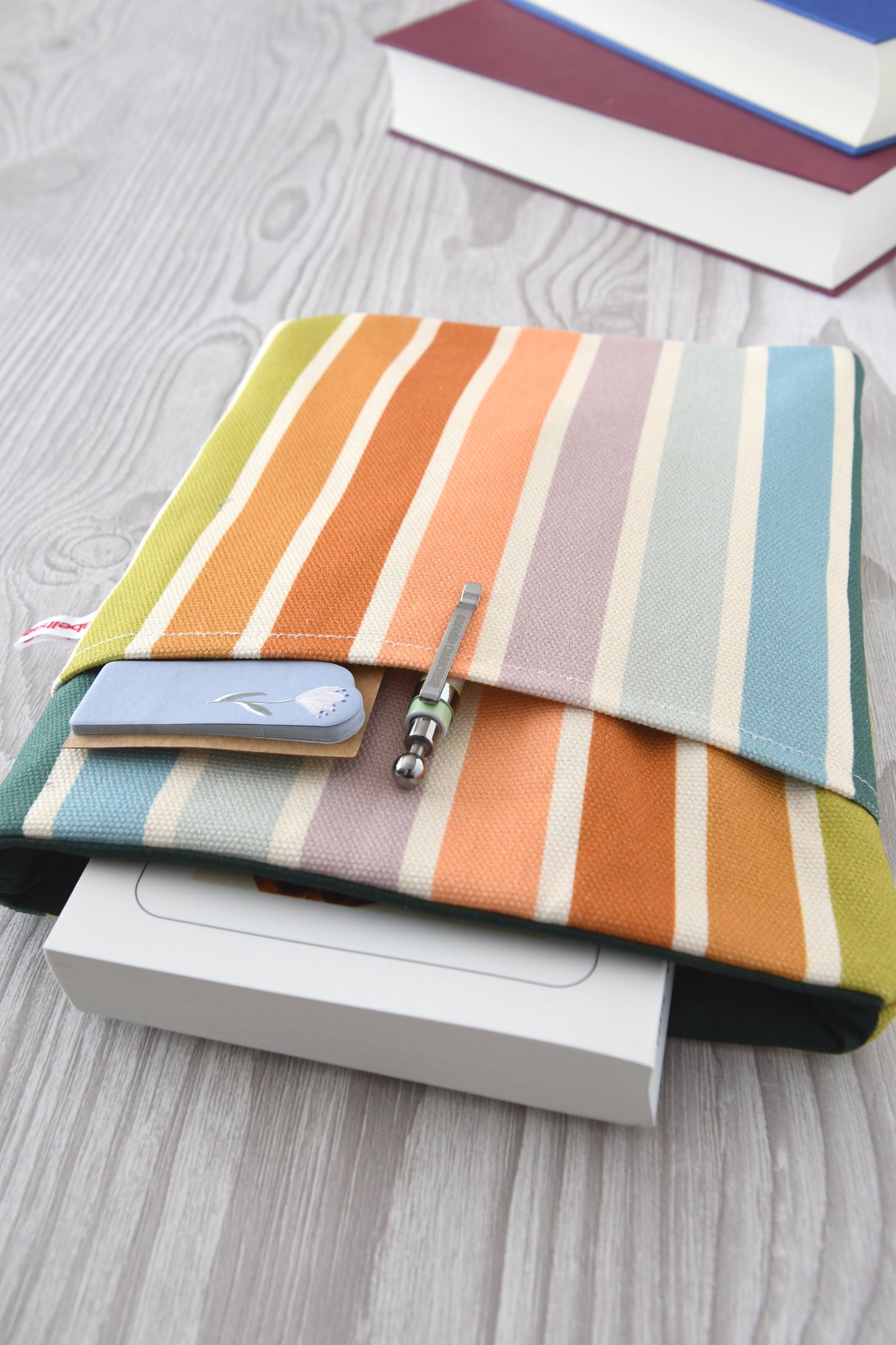 Draufsicht auf die geöffnete Buchtasche im Pastellstreifen-Design mit eingestecktem Buch sowie einem Stift und Lesezeichen in der praktischen Vordertasche.