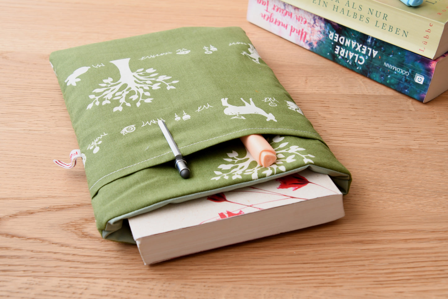 Gemütliche Buchhülle mit Waldtier-Motiv. Ein Taschenbuch ist in der weich gepolsterten Tasche verstaut, während im praktischen Extra-Fach auf der Vorderseite zwei Stifte stecken