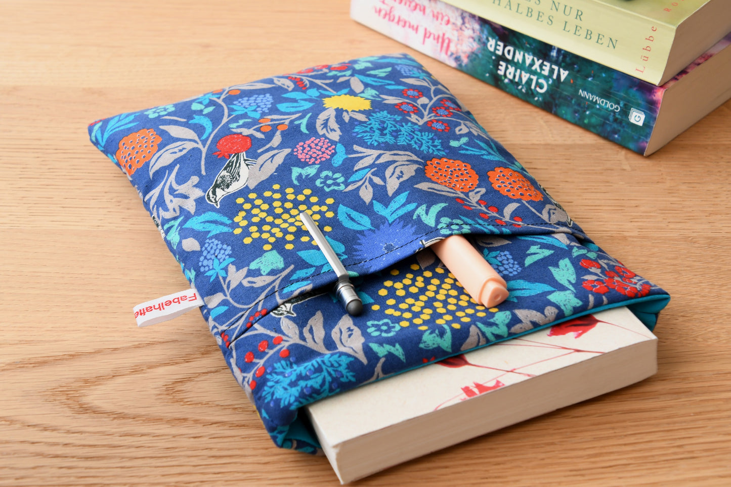 Elegante Buchhülle in Blau mit floralem Muster und kleinen Vögeln. Ein Roman schaut oben ein Stück aus der weich gepolsterten Tasche heraus, während im cleveren Zubehörfach auf der Vorderseite zwei Stifte griffbereit stecken.