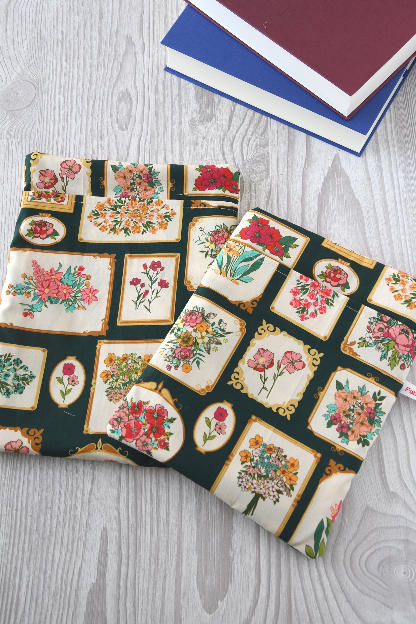 Handgemachte Buchtaschen aus Stoff im Größenvergleich: Die kleine Buchhülle (lrechts, für Taschenbücher) liegt neben der großen Schutzhülle (links, für gebundene Bücher & Tablets). Beide Taschen der Fabelhaften Werkstatt zeigen das Muster „Bildergalerie“ mit eingerahmten Blumenmotiven und sind weich gepolstert.