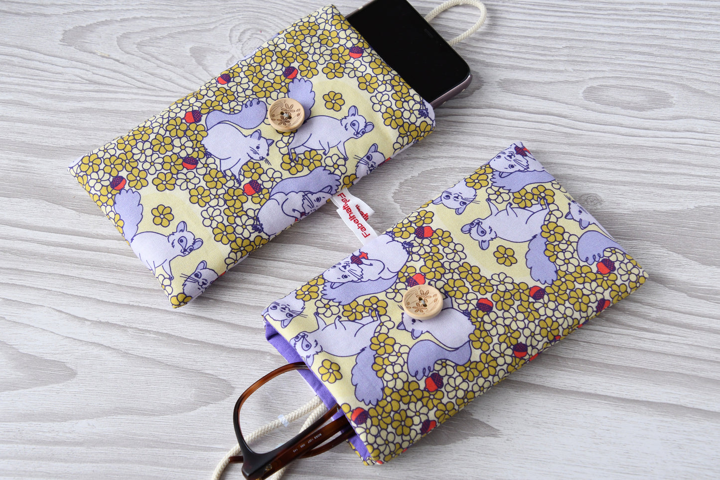 Zwei handgenähte Stofftaschen mit süßem Eichhörnchen-Muster von Fabelhafte Werkstatt nebeneinander: Eine genutzt als Handyhülle mit Smartphone, die andere als Brillenetui mit Sonnenbrille.