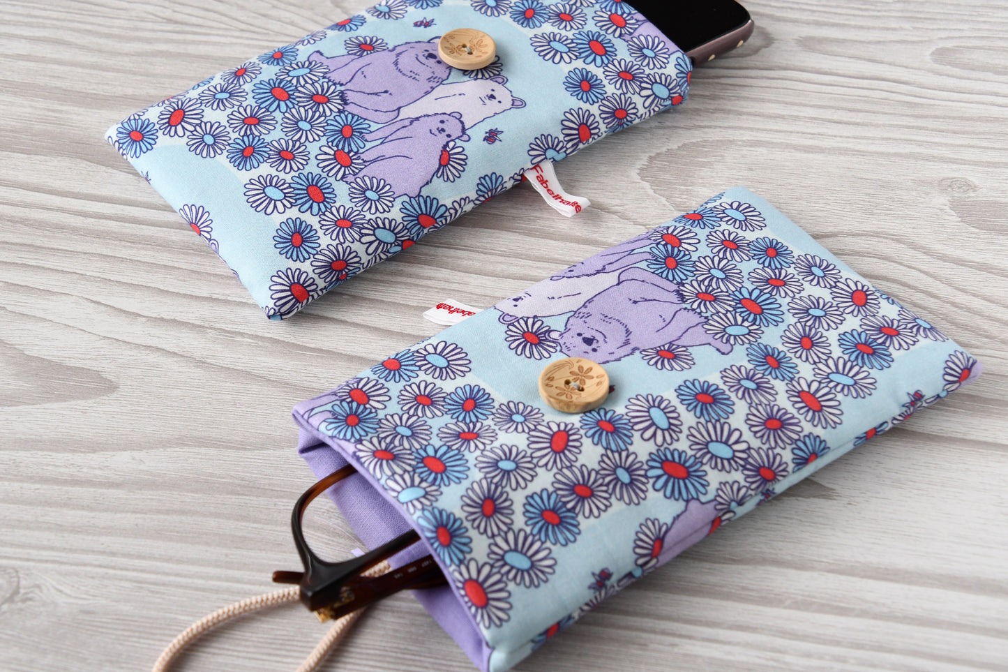 Zwei handgenähte Stofftaschen mit süßem Bären-Muster von Fabelhafte Werkstatt nebeneinander: Eine genutzt als Handyhülle mit Smartphone, die andere als Brillenetui mit Brille.