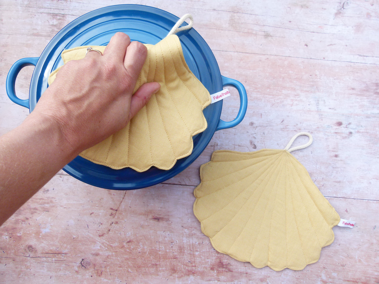 Präsentation des maritimen Topflappen-Sets an einem Kochtopf. Ein muschelförmiger Topflappen aus gelbem Leinen liegt flach auf dem Topfdeckel, während der zweite Topflappen dekorativ daneben auf der Küchenzeile platziert ist.
