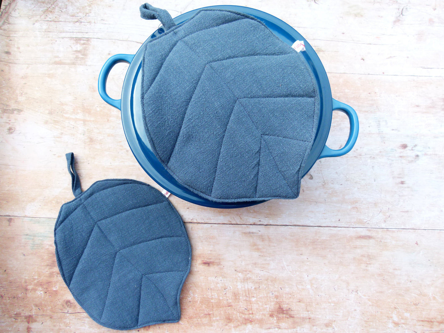 Ein Set aus zwei handgenähten, blattförmigen Topflappen aus blauem Leinen an einem Topf. Ein blauer Topflappen liegt flach auf dem Deckel des geschlossenen Topfes, während der zweite Topflappen dekorativ daneben auf der Arbeitsfläche platziert ist.