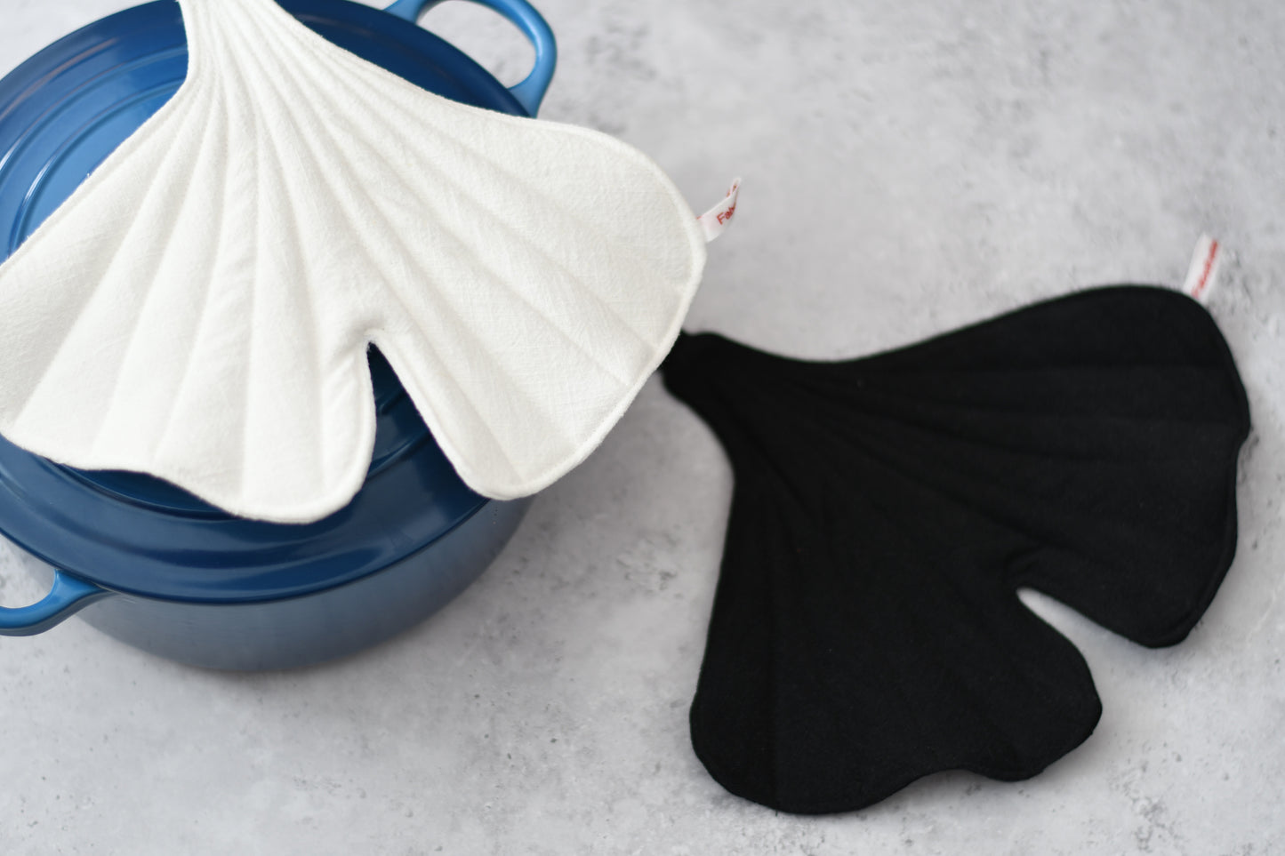 Ein Set aus zwei handgenähten, ginkgoförmigen Topflappen liegt dekorativ auf dem Rand eines Topfes. Der eine Topflappen ist aus tiefschwarzem Leinen gefertigt, der andere aus reinweißem Leinen. Beide zeigen die markante, organische Form eines Ginkgoblattes mit feiner Steppung und Aufhängeschlaufe.