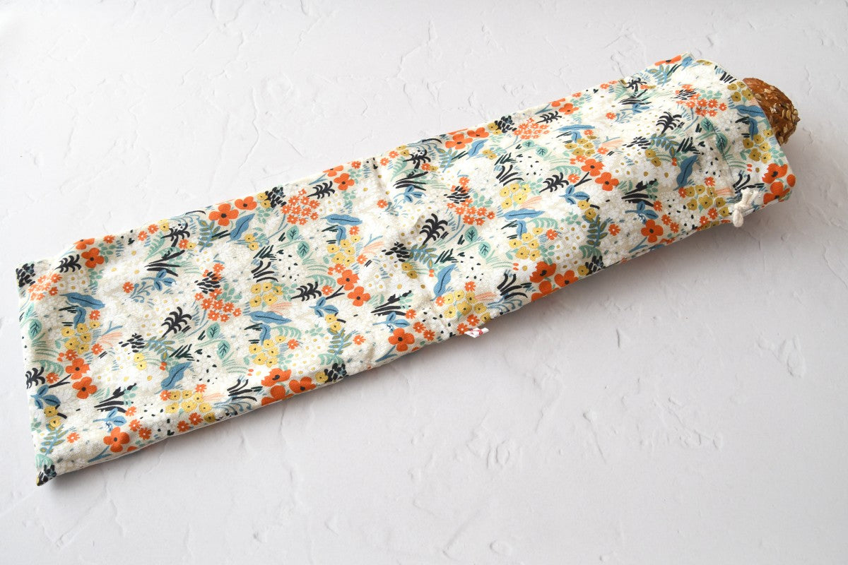 Ein handgenähter, langer Baguettebeutel aus  liegt auf einer hellen Küchenarbeitsplatte. Ein frisches, knuspriges Baguette schaut oben leicht aus der geöffneten Kordelöffnung heraus.