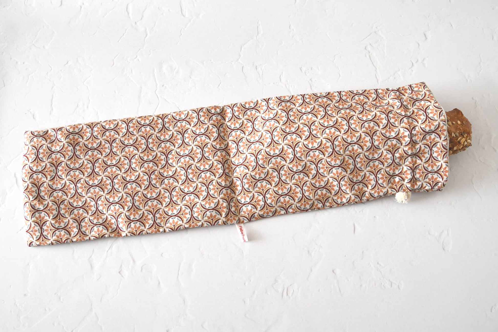 Ein handgenähter, langer Baguettebeutel liegt auf einer hellen Küchenarbeitsplatte. Ein frisches, knuspriges Baguette schaut oben leicht aus der geöffneten Kordelöffnung heraus.