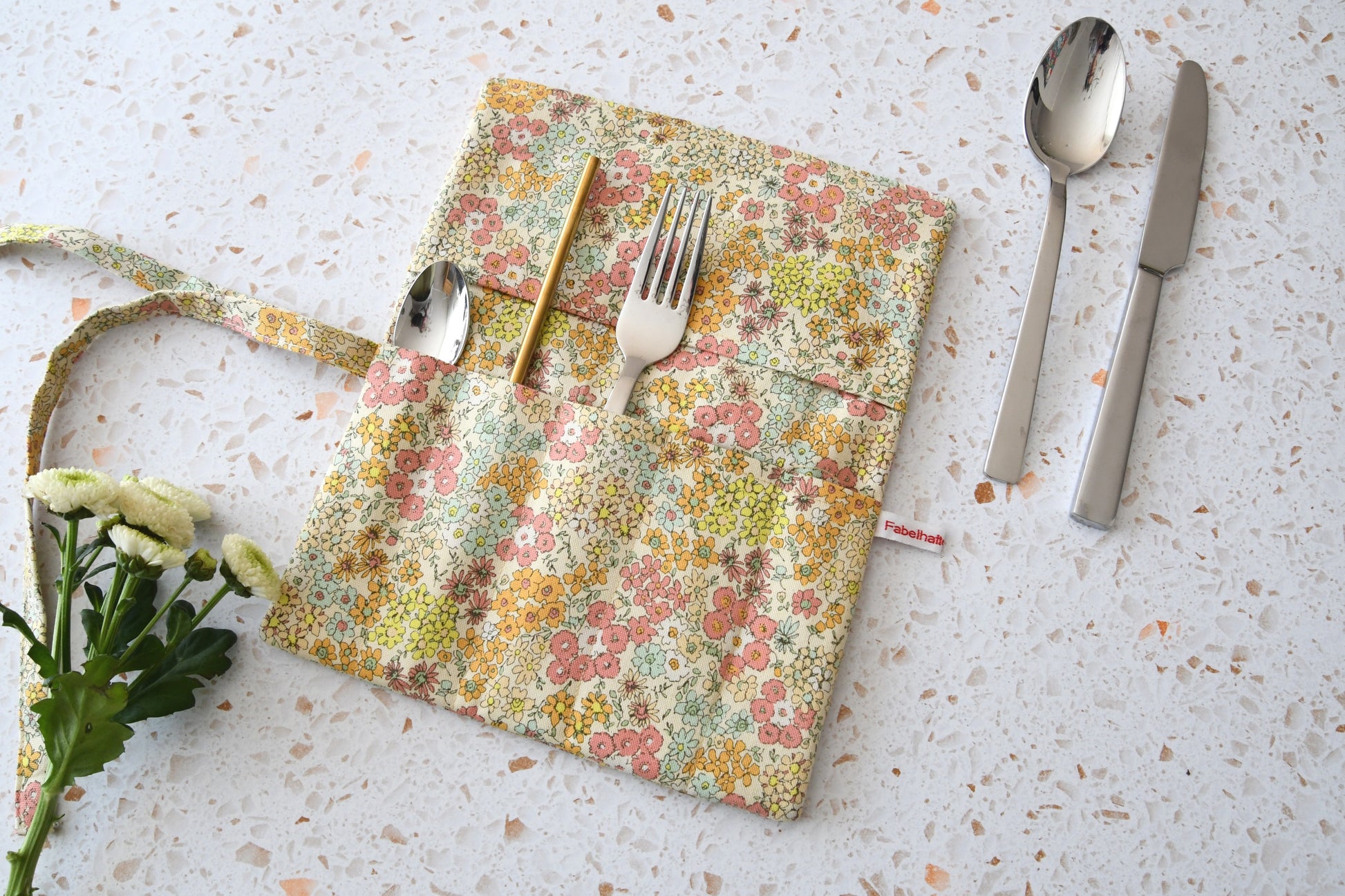 Handgenähte Besteckrolle aus Stoff mit romantischem Blumenmuster, geöffnet und mit Strohhalm, Gabel und Löffel in den Fächern bestückt.