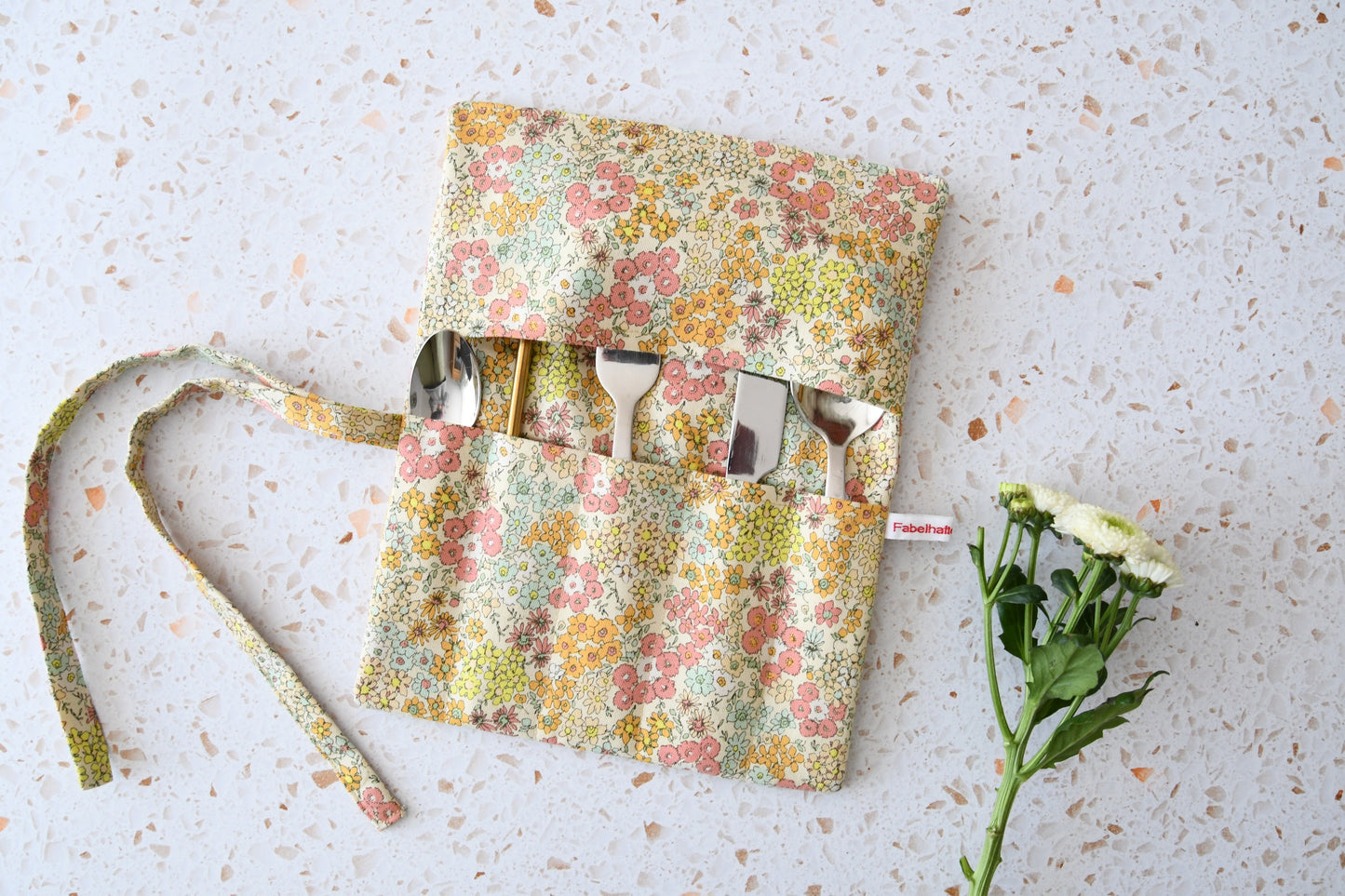 Geöffnete Besteckrolle aus Stoff mit romantischem Blumenmuster, bestückt mit Messer, Gabel und Löffel in den Einsteckfächern.