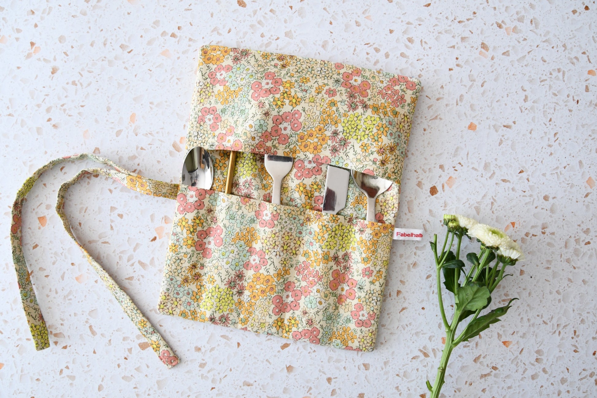 Geöffnete Besteckrolle aus Stoff mit romantischem Blumenmuster, bestückt mit Messer, Gabel und Löffel in den Einsteckfächern.
