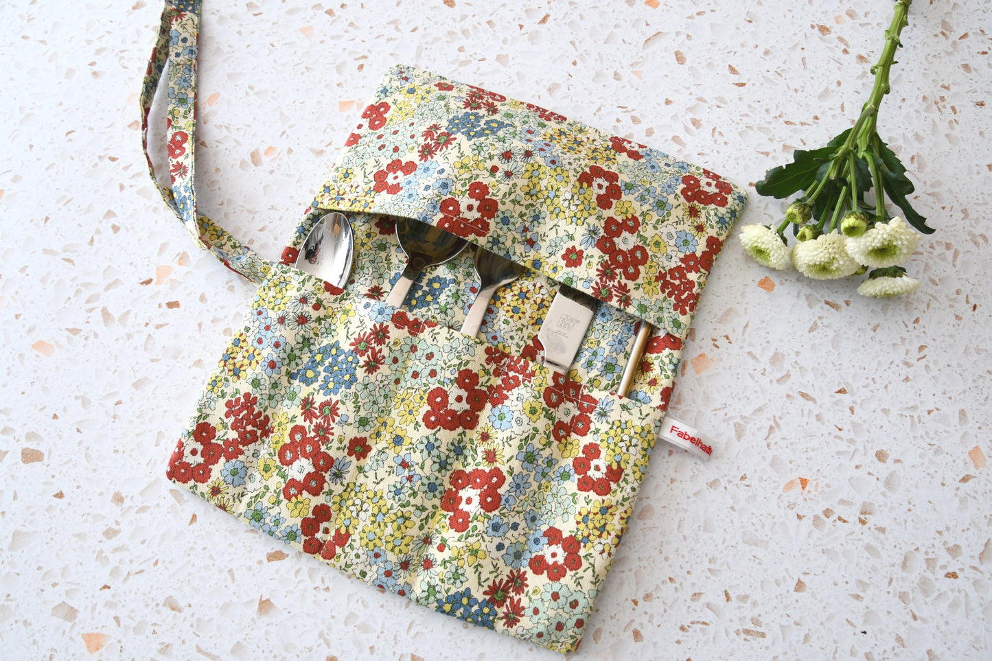 Handgenähte Besteckrolle aus Stoff mit zauerhaftem Blumenmuster, geöffnet und mit Messer, Gabel und Löffel in den Fächern bestückt.