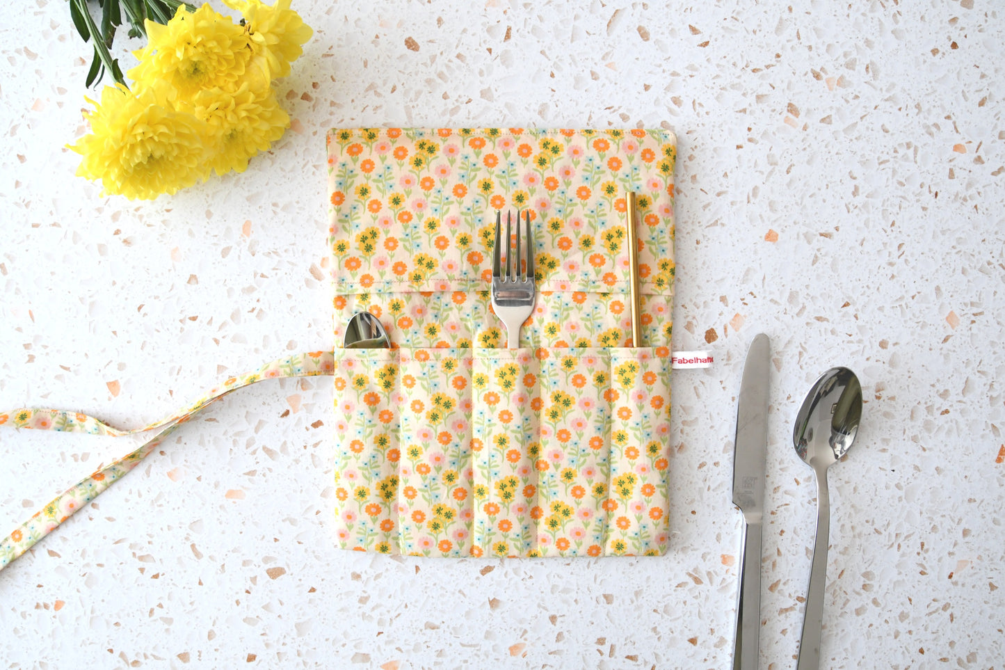 Handgenähte Besteckrolle aus Stoff mit sommerlichem Blumenmuster, geöffnet und mit Messer, Gabel und Löffel in den Fächern bestückt.