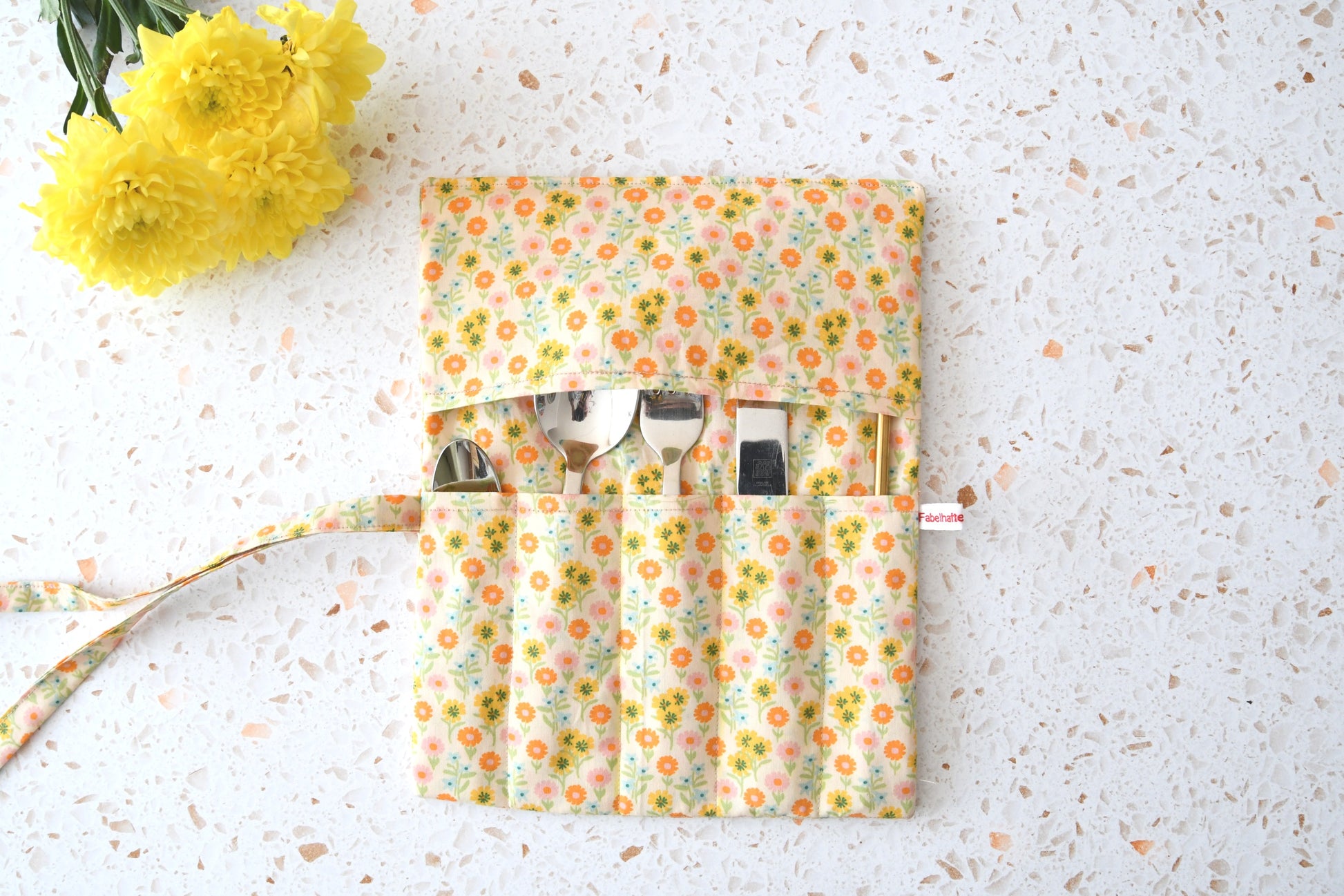 Handgenähte Besteckrolle aus Stoff mit sommerlichem Blumenmuster, geöffnet und mit Messer, Gabel und Löffel in den Fächern bestückt.
