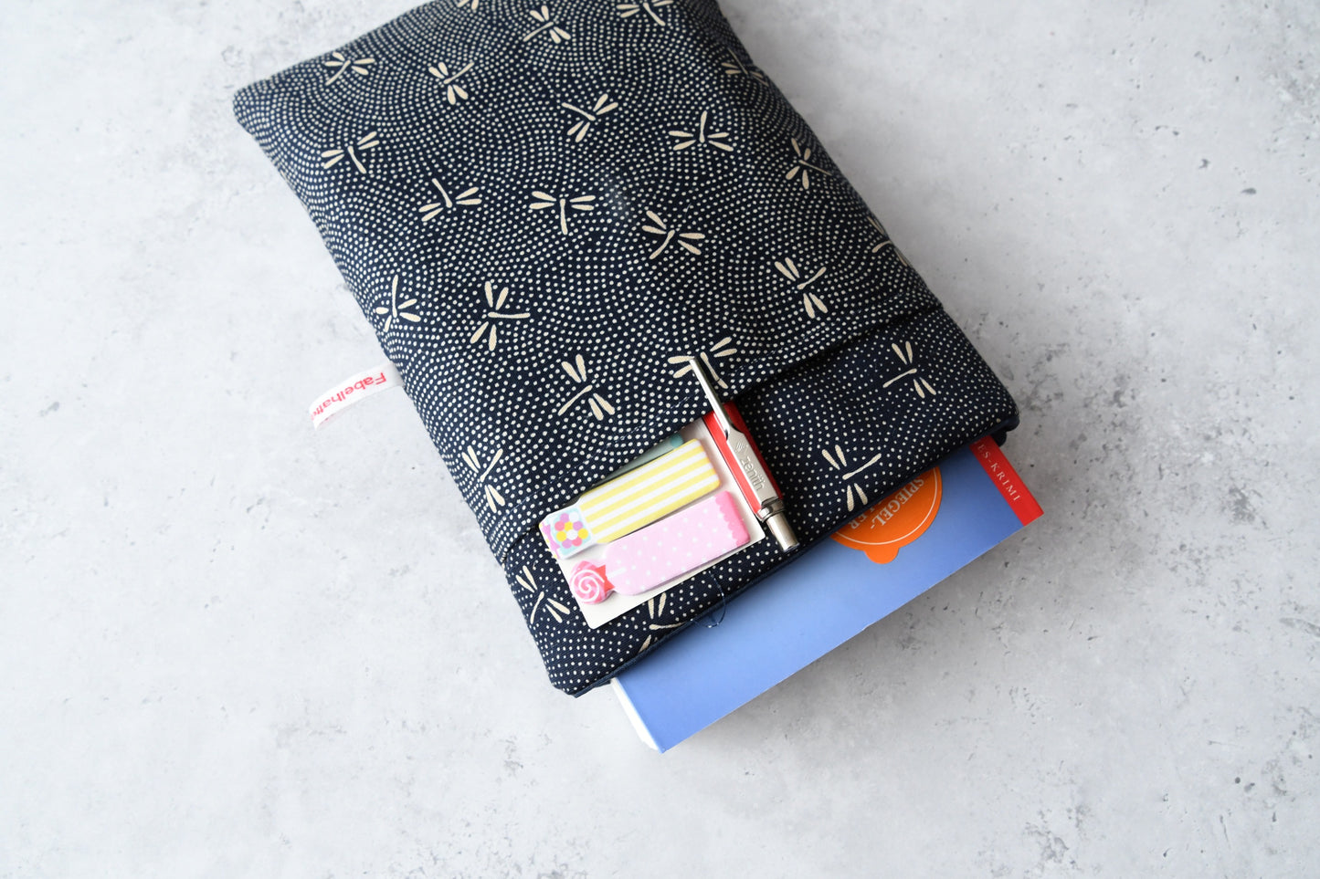 Elegante dunkelblaue Buchhülle mit hellen japanischen Libellen-Motiven in Benutzung. Ein Taschenbuch schaut oben leicht aus der weich gefütterten Hülle heraus, während im cleveren Vorderfach ein Stift und ein Textmarker stecken.