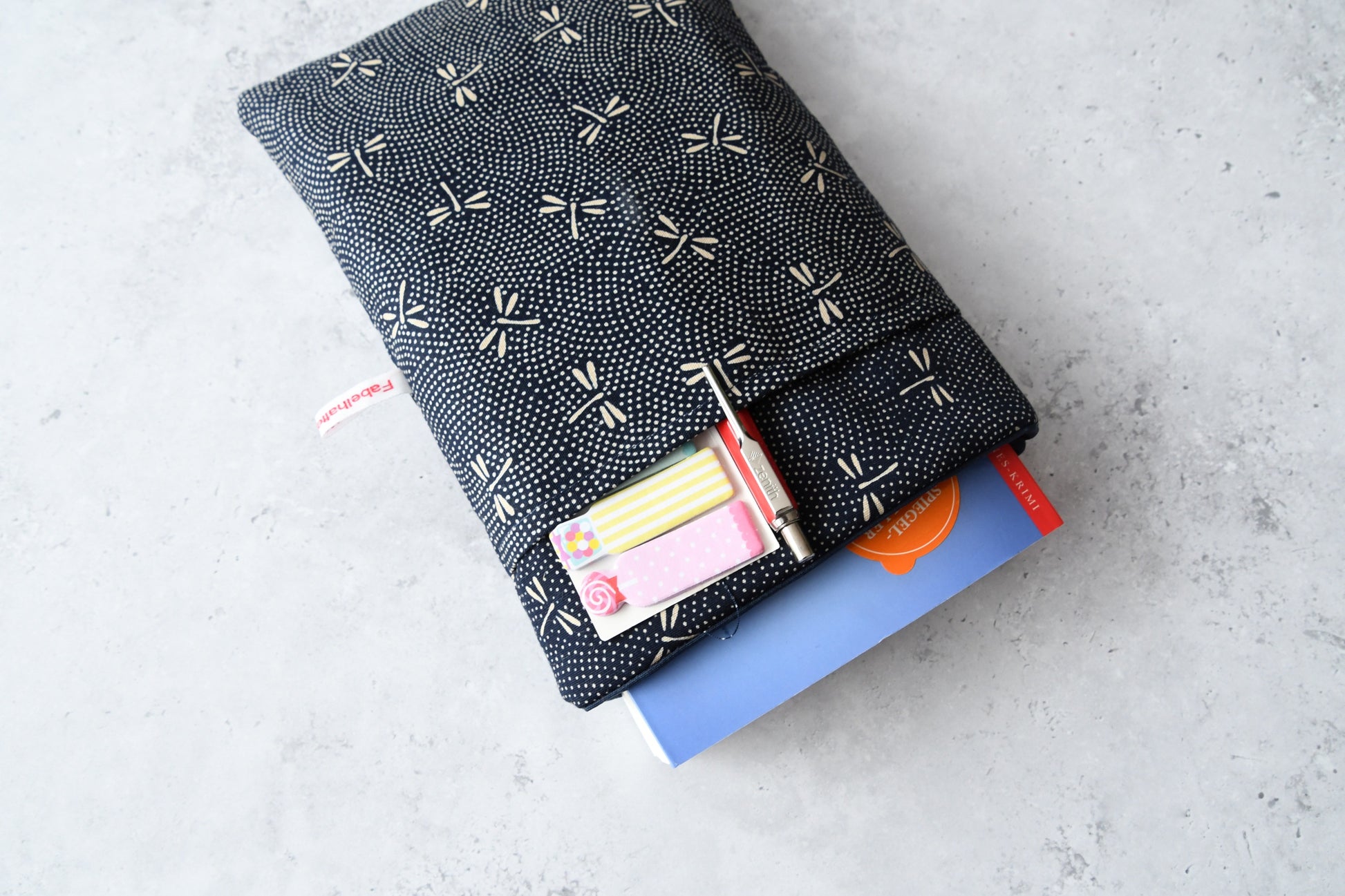 Elegante dunkelblaue Buchhülle mit hellen japanischen Libellen-Motiven in Benutzung. Ein Taschenbuch schaut oben leicht aus der weich gefütterten Hülle heraus, während im cleveren Vorderfach ein Stift und ein Textmarker stecken.