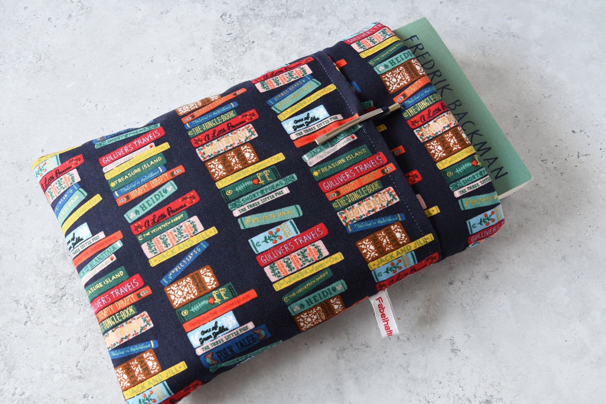 Detailreiches Foto einer Buchtasche mit Bücherrücken-Print. Aus der weichen Hülle schaut ein echtes Buch heraus. Das clevere Zubehörfach auf der Vorderseite ist mit einem Kugelschreiber bestückt, um den perfekten Schutz für unterwegs zu demonstrieren.