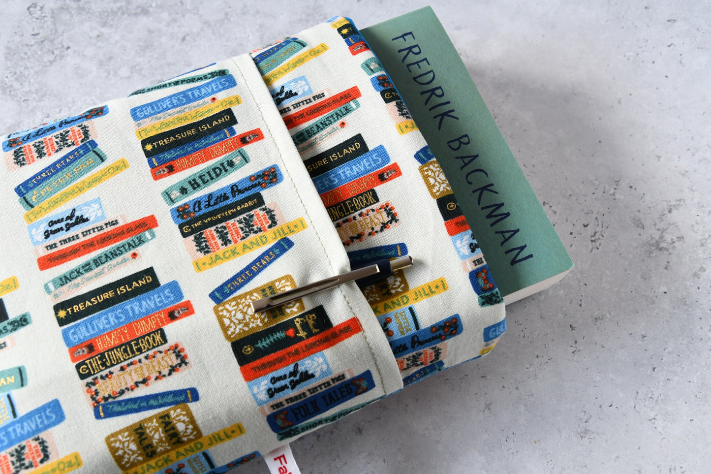 Detailreiches Foto einer Buchtasche mit Bücherrücken-Print. Aus der weichen Hülle schaut ein echtes Buch heraus. Das clevere Zubehörfach auf der Vorderseite ist mit einem Stift bestückt, um den perfekten Schutz für unterwegs zu demonstrieren.