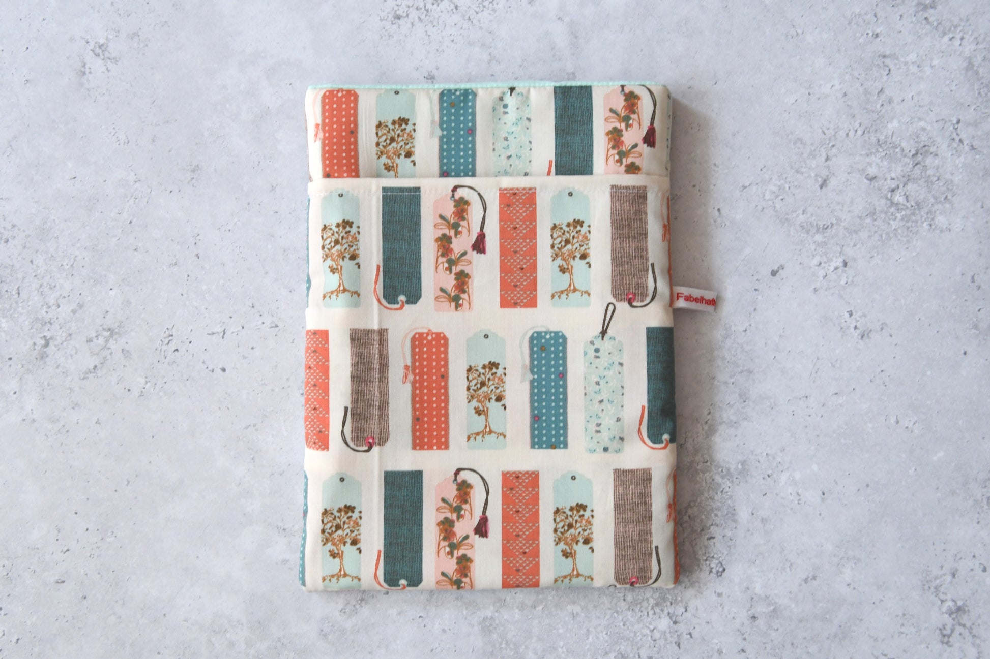 Handgefertigte, gepolsterte Buchtasche der Fabelhaften Werkstatt aus hellem Stoff, bedruckt mit einem kreativen Muster aus vielen bunten, unterschiedlich gestalteten Lesezeichen. Das Produktfoto zeigt das flach liegende Design mit dem praktischen Vorderfach.