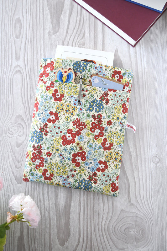 Handgefertigte, weich gepolsterte Buchtasche der Fabelhaften Werkstatt aus hellem Stoff mit einem zarten, detailreichen Streublumen-Muster in Pastelltönen. Gut sichtbar ist das aufgesetzte Vorderfach für Zubehör und die hochwertige Verarbeitung.