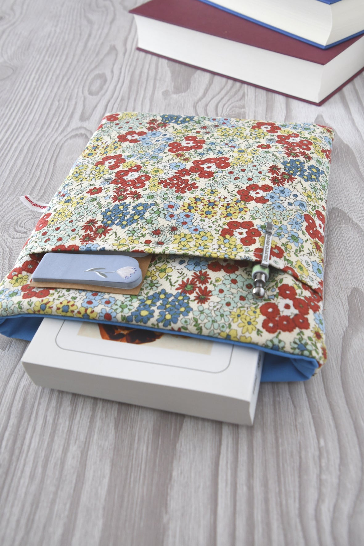  Buchtasche mit einem zarten, bunten Streublumen-Muster auf hellem Stoff. Ein Taschenbuch schaut oben ein Stück aus der weich gepolsterten Hülle heraus. Im vorderen Fach stecken ordentlich zwei Stifte für die nächste Lesepause.