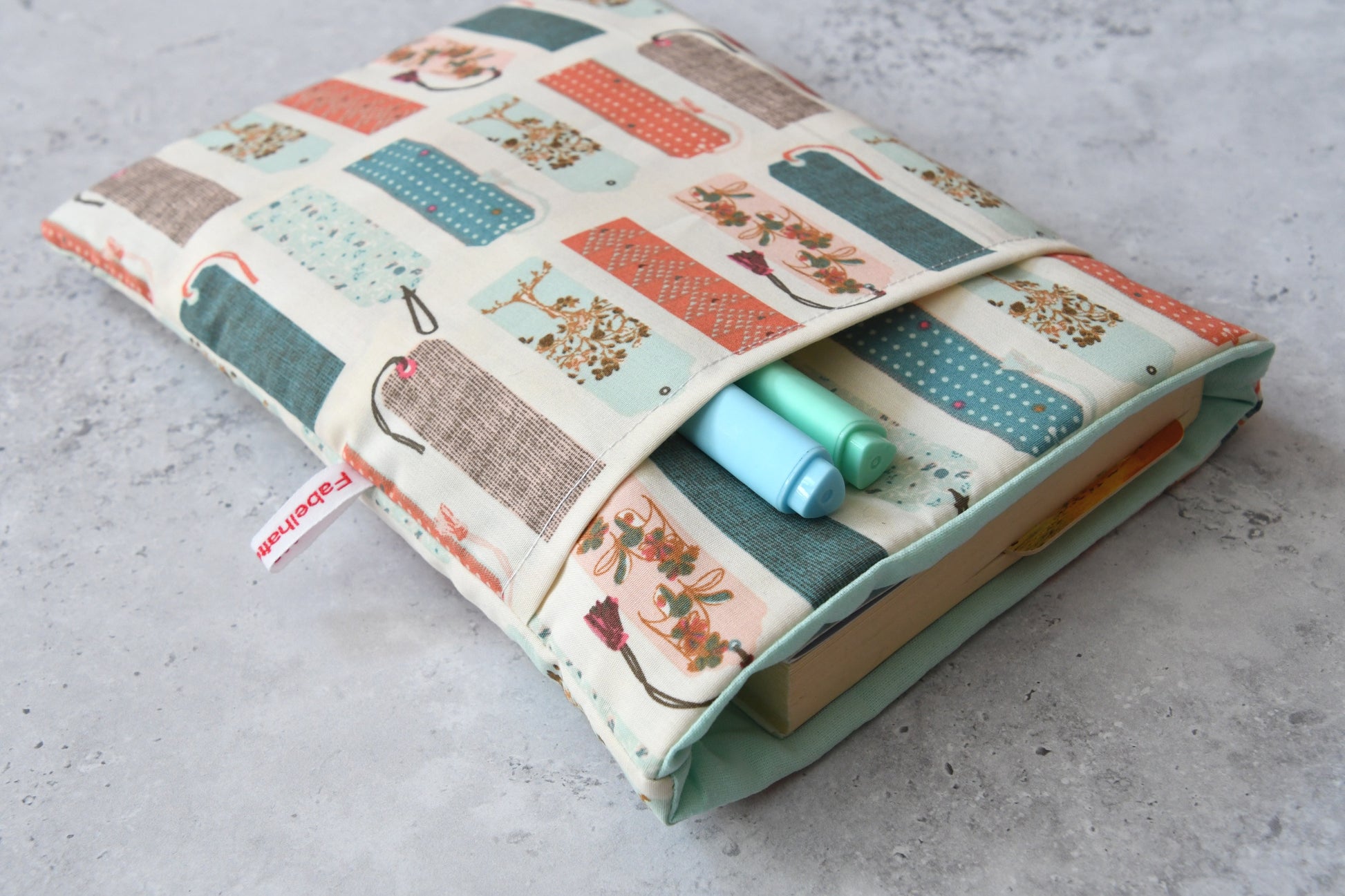 Fröhliche Buchhülle mit buntem Lesezeichen-Motiv in Benutzung. Ein Taschenbuch schaut oben ein Stück aus der weich gefütterten Tasche heraus, während im cleveren Zubehörfach auf der Vorderseite zwei Stifte griffbereit platziert sind.