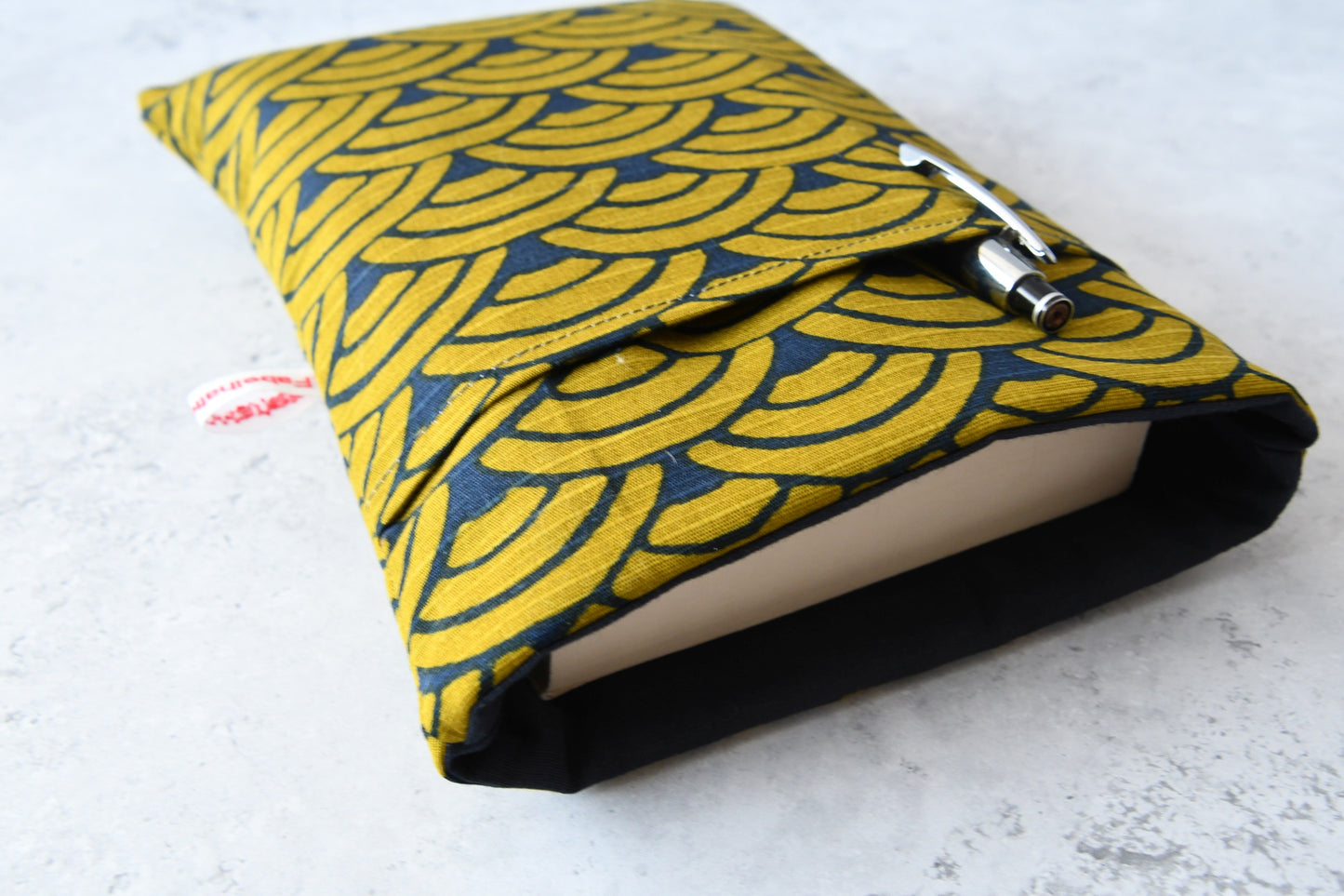 Elegante, dunkelblaue Buchtasche mit goldenem japanischen Wellenmuster in Benutzung. Ein dickes Taschenbuch schaut oben leicht aus der gepolsterten Hülle heraus. Im vorderen Fach steckt ein Kugelschreiber.