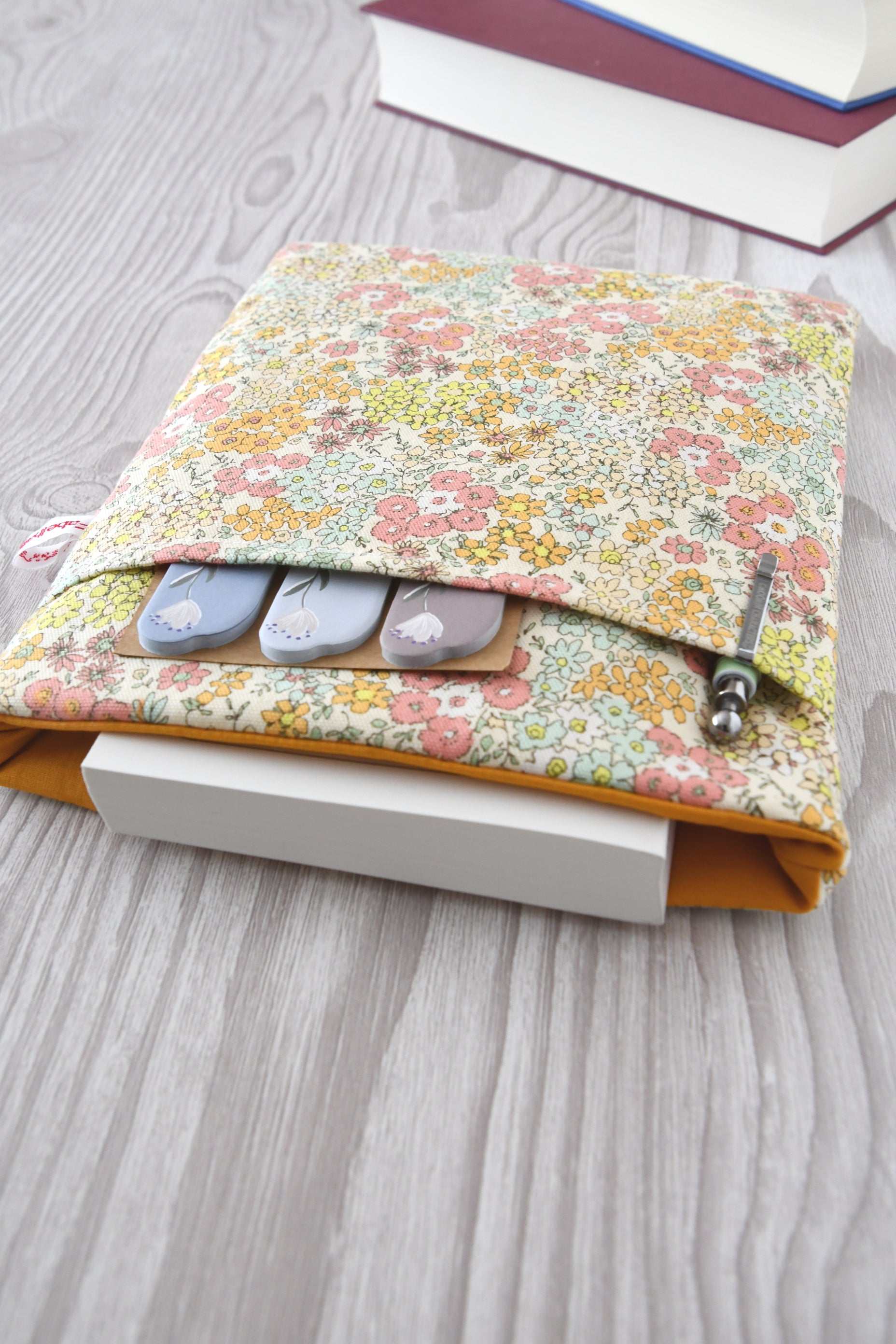 Draufsicht auf die geöffnete Blumen-Buchtasche mit eingestecktem Buch sowie einem Stift und Lesezeichen in der praktischen Vordertasche.