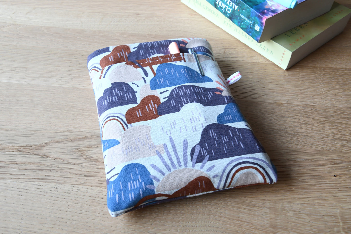 Ein Foto einer handgenähten Buchhülle. Das Muster zeigt fröhliche, bunte Wolken in Blau, Lila und Braun sowie Sonnen auf einem hellen Hintergrund. Die Buchhülle ist weich gepolstert und hat ein praktisches Fach auf der Vorderseite.