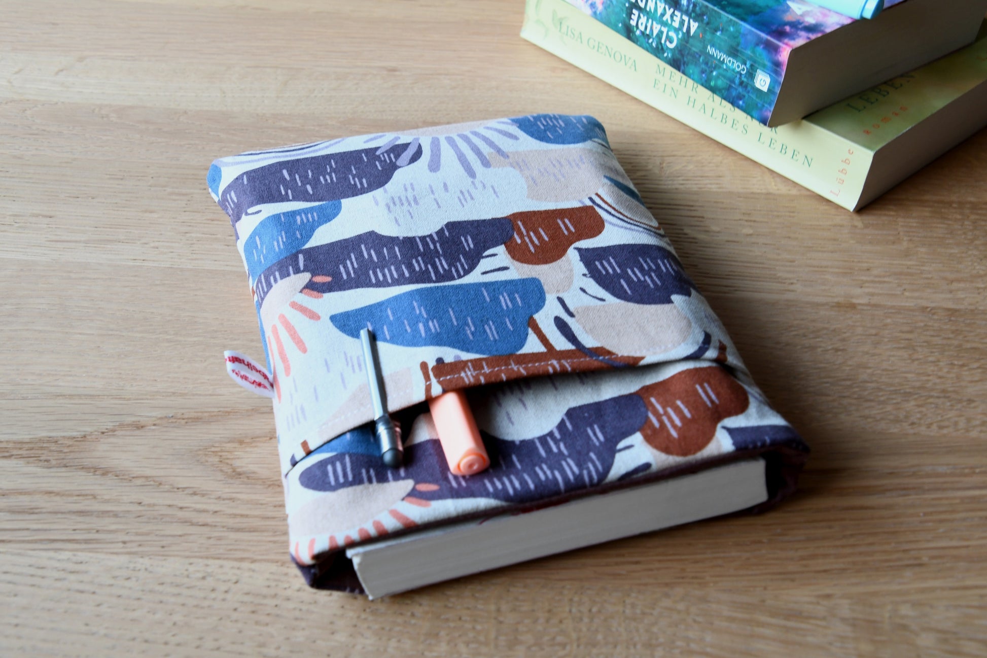 Fröhliche, bunt gemusterte Wolken-Buchtasche mit Sonne, fotografiert mit Inhalt. Aus der oberen Öffnung ragt ein Roman heraus. Das praktische Vorderfach ist mit bunten Finelinern und einem Ladekabel befüllt, um die Funktion als Alltagsbegleiter zu zeigen.