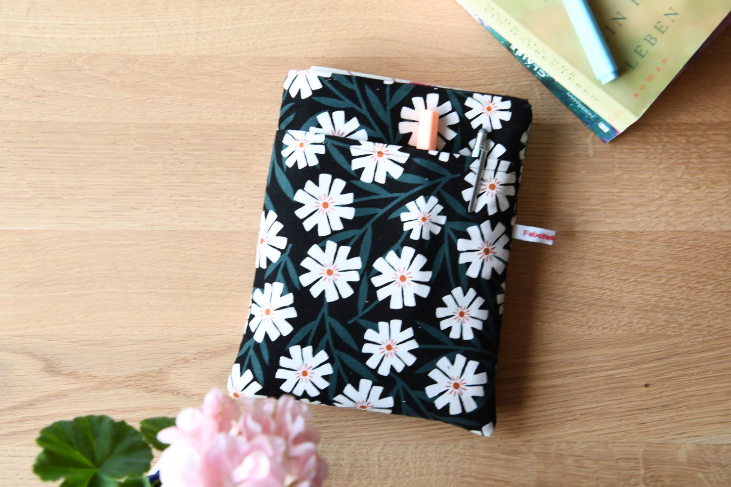 Fröhliche, weich gepolsterte Buchtasche mit einem sommerlichen Margeriten-Muster (weiße Blüten mit orangenfarbender Mitte).  Im cleveren Vorderfach sind Stifte und kleine Haftnotizen griffbereit platziert.