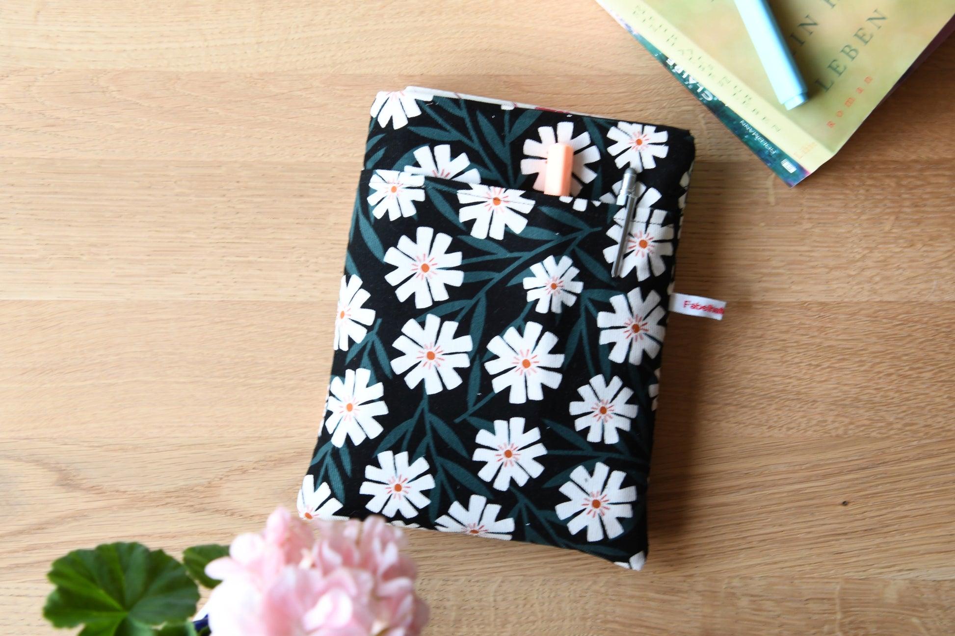 Fröhliche, weich gepolsterte Buchtasche mit einem sommerlichen Margeriten-Muster (weiße Blüten mit orangenfarbender Mitte).  Im cleveren Vorderfach sind Stifte und kleine Haftnotizen griffbereit platziert.