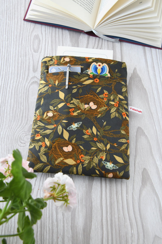 Handgefertigte, gepolsterte Buchtasche der Fabelhaften Werkstatt aus braunem Stoff mit einem detailreichen Natur-Motiv aus kleinen Vogelnestern und hellen Vogeleiern. Gut sichtbar ist das aufgesetzte Vorderfach für Zubehör auf der flach liegenden Hülle.