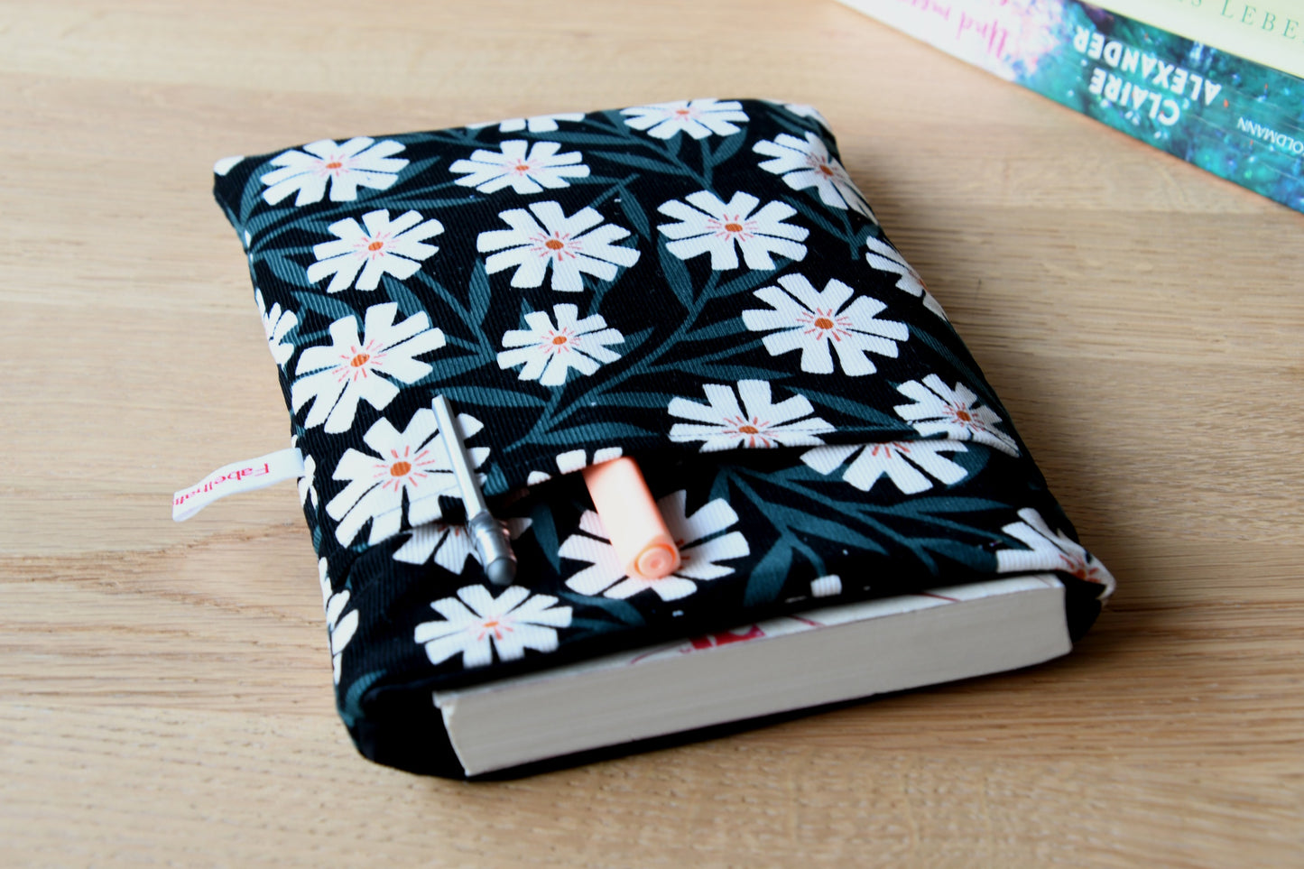 Fröhliche, weich gepolsterte Buchtasche mit einem sommerlichen Margeriten-Muster (weiße Blüten mit orangenfarbender Mitte). Aus der oberen Öffnung ragt ein Roman heraus, und im Vorderfach sind Stifte und kleine Haftnotizen griffbereit platziert.
