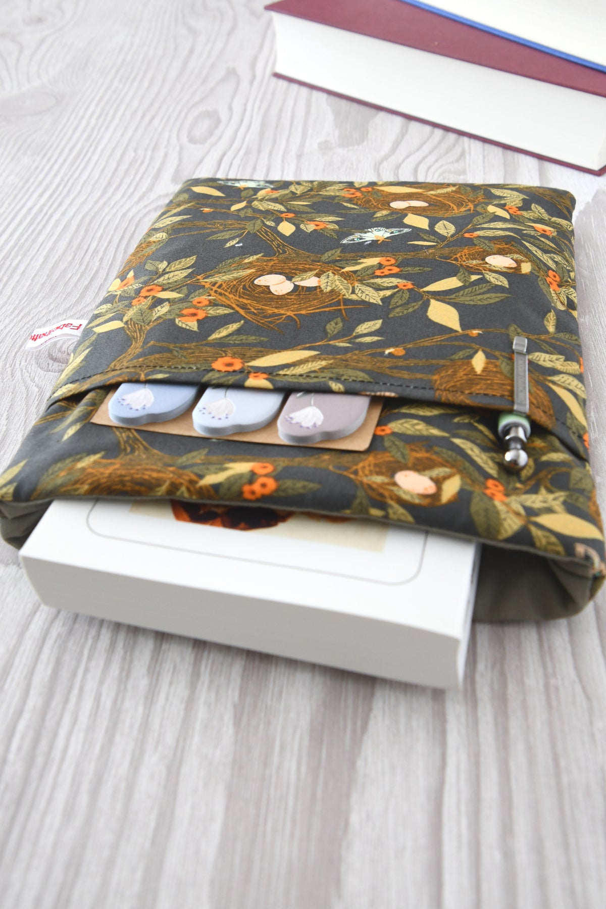 Gemütliche Buchhülle im Natur-Look mit Vogelnest-Muster auf braunem Grund. Ein dickes Taschenbuch schaut oben leicht aus der weich gepolsterten Tasche heraus. Im praktischen Vorderfach stecken ordentlich zwei Schreibstifte.
