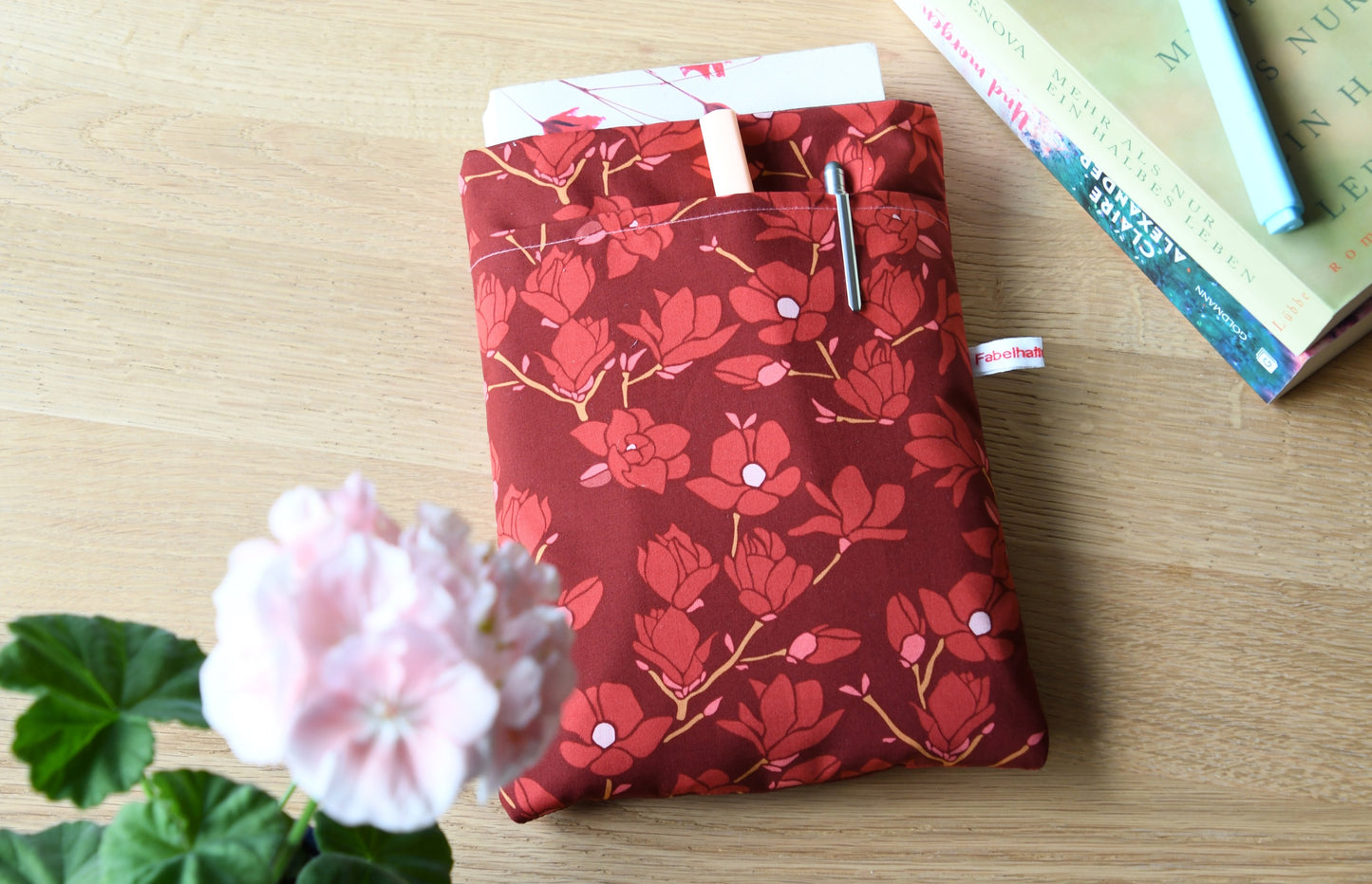 Elegante Buchhülle aus edlem Stoff mit wunderschönen Magnolien-Blüten. Das Produktfoto zeigt die flach liegende, gepolsterte Tasche mit Fokus auf das clevere Zubehörfach und die sauberen Nähte.