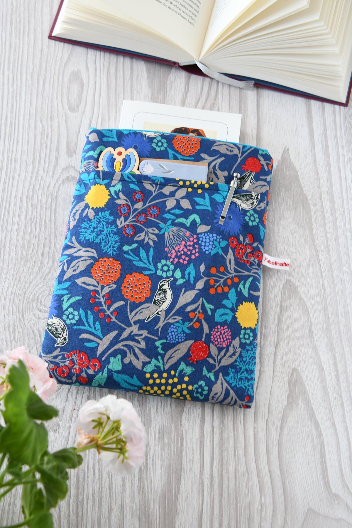 Handgefertigte, gepolsterte Buchtasche der Fabelhaften Werkstatt aus blauem Stoff, bedruckt mit einem eleganten Blumenmuster und kleinen Vögeln zwischen den Blüten. Das Foto zeigt das praktische, aufgesetzte Vorderfach auf der flach liegenden Hülle.