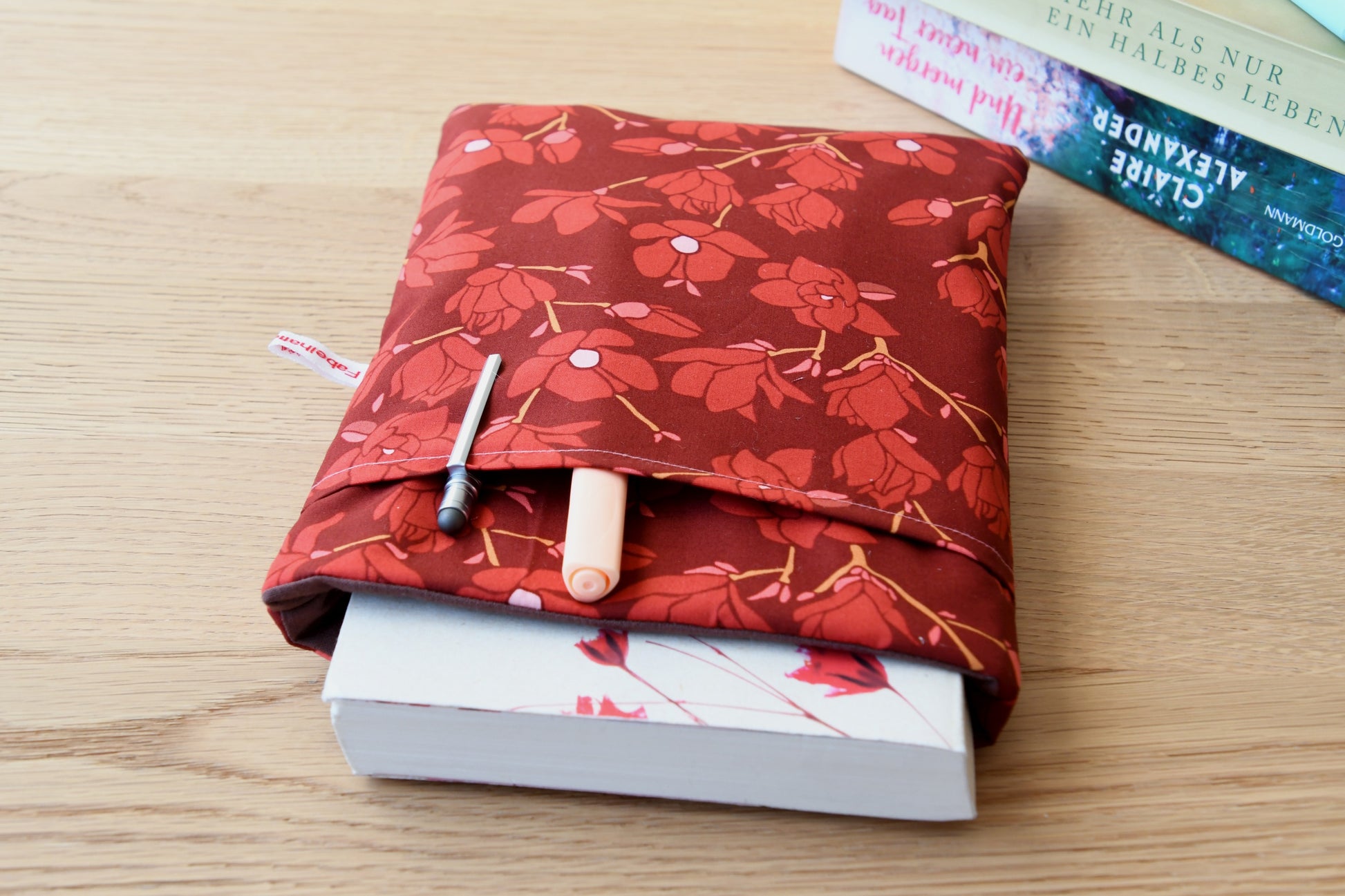Elegante, handgefertigte Buchhülle mit edlen Magnolien-Blüten. Ein dickes Buch ist sicher in der Tasche verstaut, während im praktischen Zubehörfach auf der Vorderseite ein Textmarker und Fineliner stecken.