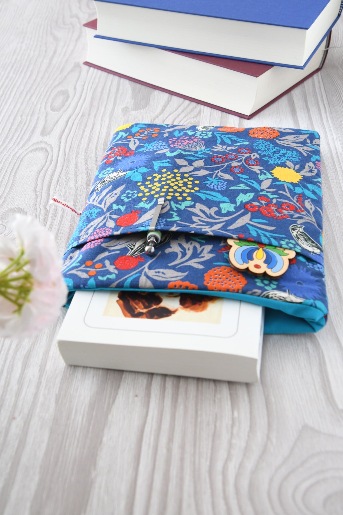 Elegante Buchhülle in Blau mit floralem Muster und kleinen Vögeln. Ein Roman schaut oben ein Stück aus der weich gepolsterten Tasche heraus, während im cleveren Zubehörfach auf der Vorderseite zwei Stifte griffbereit stecken.