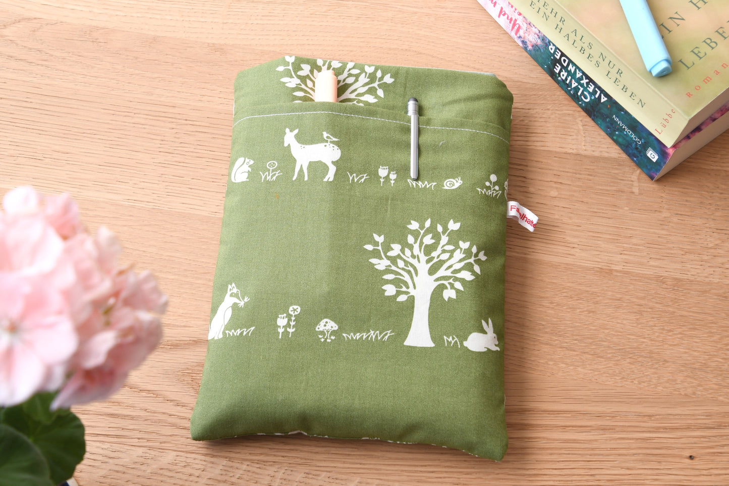Gepolsterte Buchtasche mit einem gemütlichen Wald-Motiv. Auf dem grünen Stoff sind kleine Tiere und Bäume zu sehen. Die Tasche zeigt das eingenähte „Fabelhafte Werkstatt“-Logo.