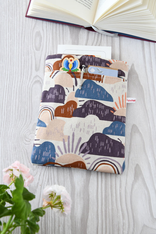 Ein Foto einer handgenähten Buchhülle. Das Muster zeigt fröhliche, bunte Wolken in Blau, Lila und Braun sowie Sonnen auf einem hellen Hintergrund. Die Buchhülle ist weich gepolstert und hat ein praktisches Fach auf der Vorderseite.