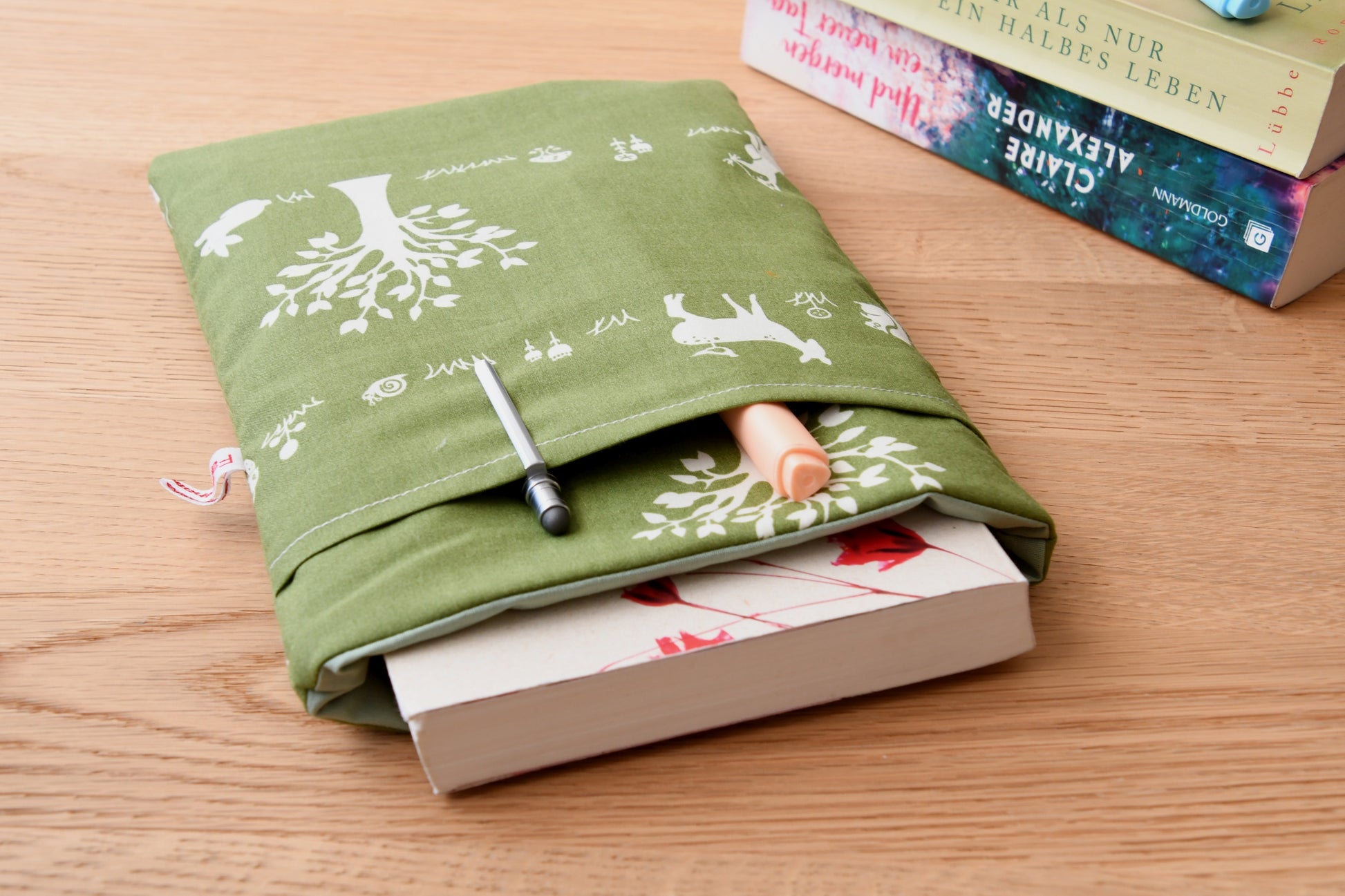 Gemütliche Buchhülle mit Waldtier-Motiv. Ein Taschenbuch ist in der weich gepolsterten Tasche verstaut, während im praktischen Extra-Fach auf der Vorderseite zwei Stifte stecken