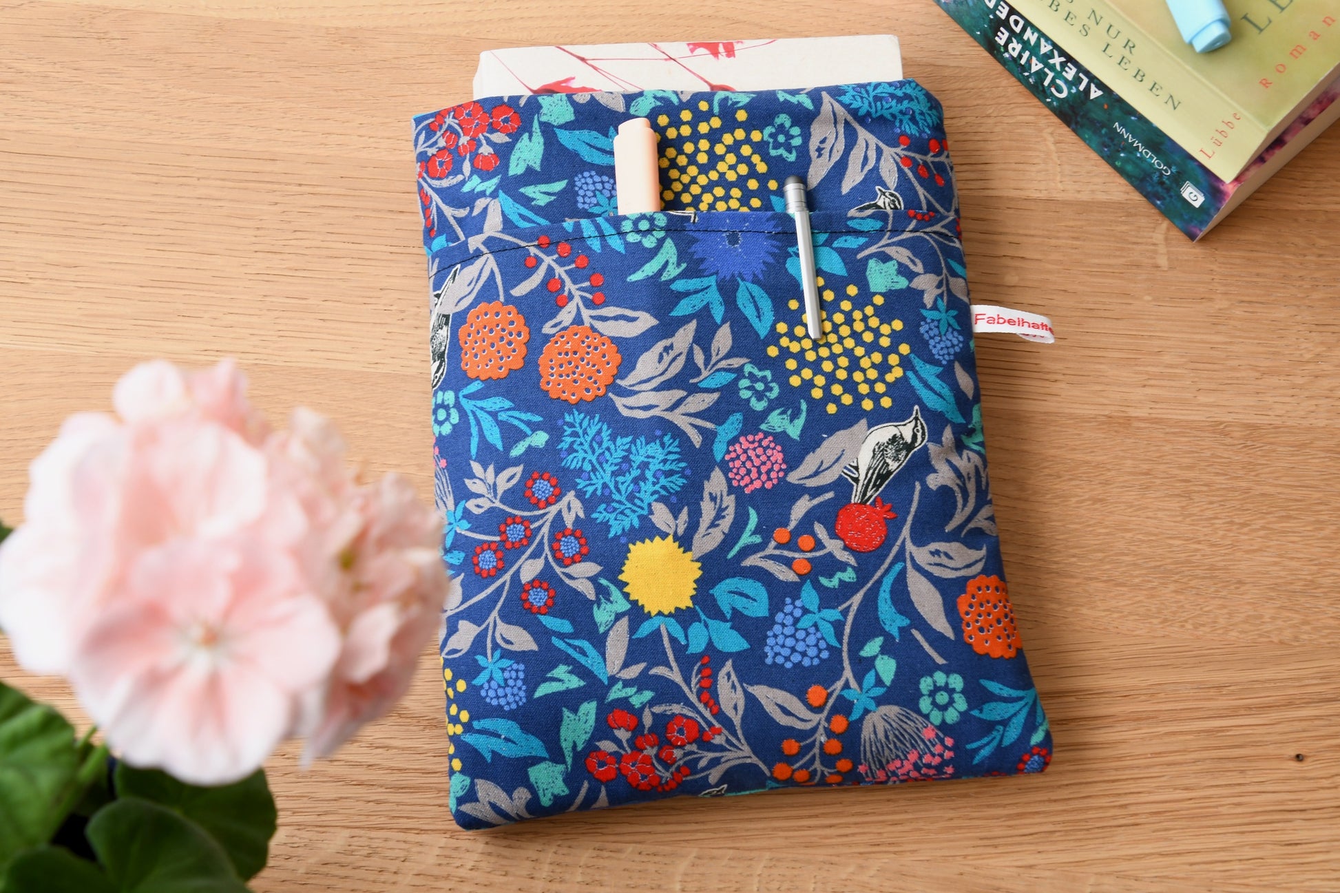 Handgefertigte, gepolsterte Buchtasche der Fabelhaften Werkstatt aus blauem Stoff, bedruckt mit einem eleganten Blumenmuster und kleinen Vögeln zwischen den Blüten. Das Foto zeigt das praktische, aufgesetzte Vorderfach auf der flach liegenden Hülle.