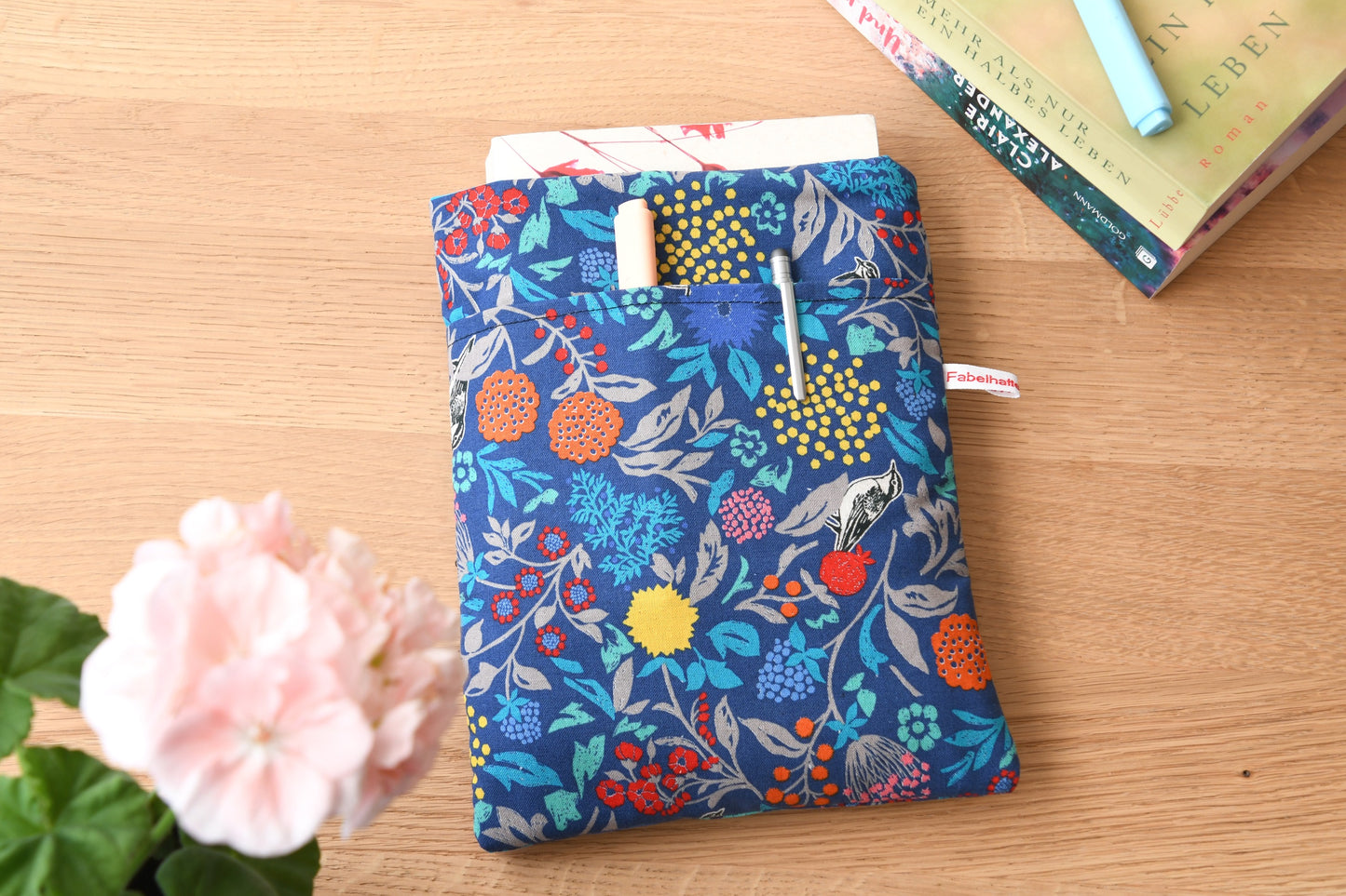 Handgefertigte, gepolsterte Buchtasche der Fabelhaften Werkstatt aus blauem Stoff, bedruckt mit einem eleganten Blumenmuster und kleinen Vögeln zwischen den Blüten. Das Foto zeigt das praktische, aufgesetzte Vorderfach auf der flach liegenden Hülle.