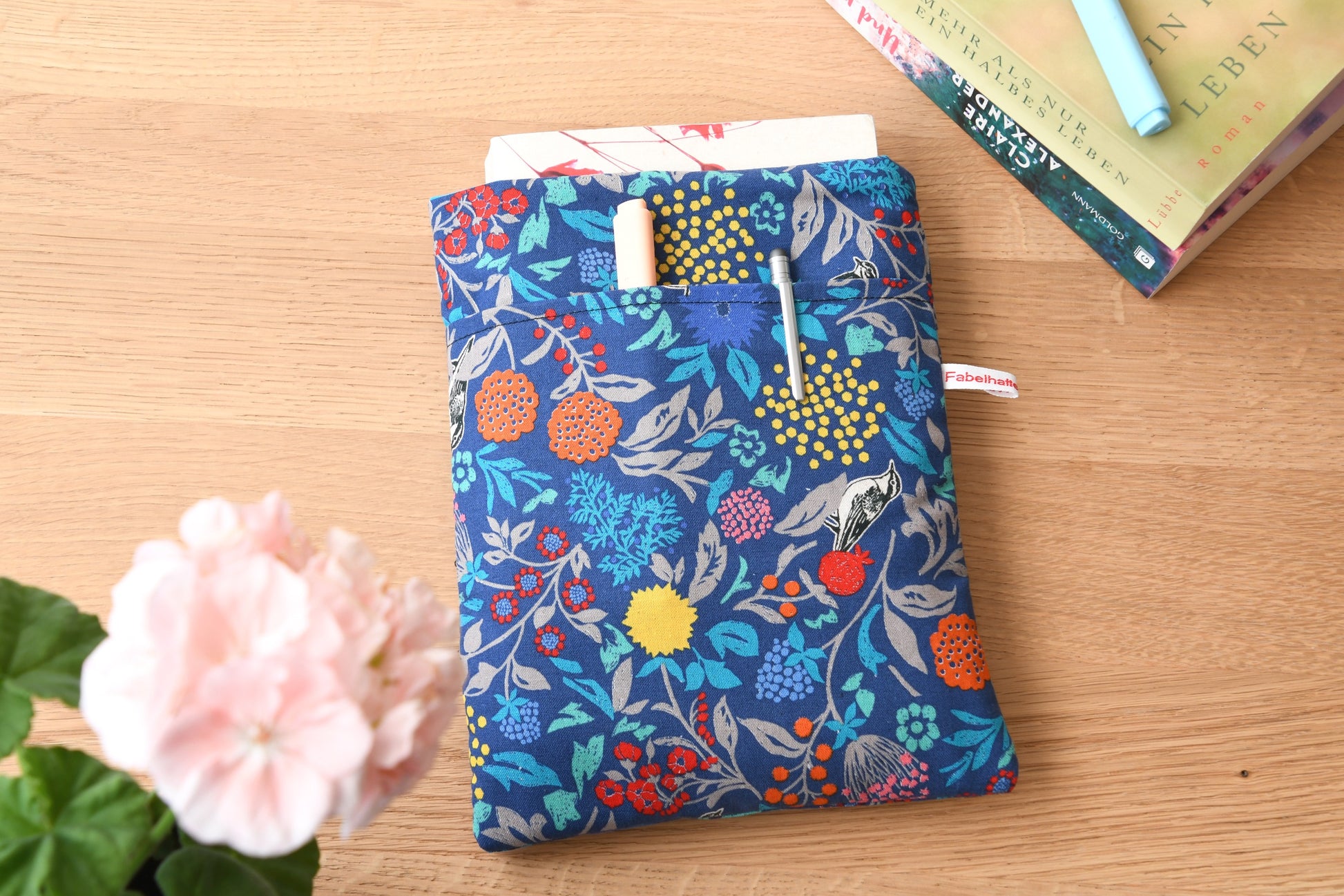 Handgefertigte, gepolsterte Buchtasche der Fabelhaften Werkstatt aus blauem Stoff, bedruckt mit einem eleganten Blumenmuster und kleinen Vögeln zwischen den Blüten. Das Foto zeigt das praktische, aufgesetzte Vorderfach auf der flach liegenden Hülle.