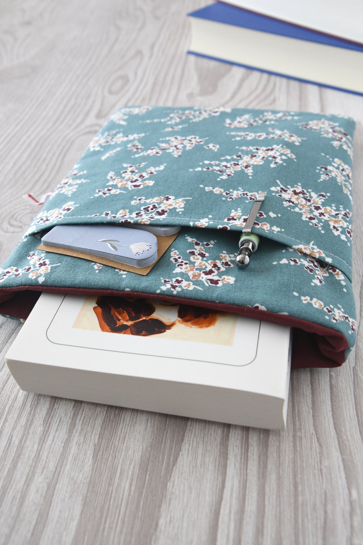 Handgefertigte, gepolsterte Buchtasche der Fabelhaften Werkstatt mit einem eleganten Muster aus herabhängenden Wisteria-Zweigen (Blauregen). Auf der flach liegenden Hülle ist das praktische, aufgesetzte Vorderfach gut zu erkennen.