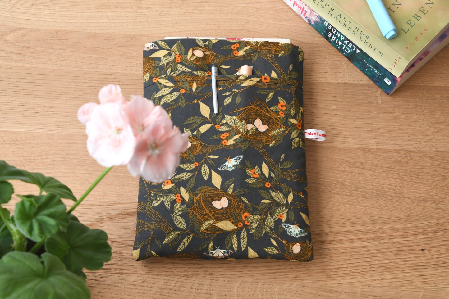 Handgefertigte, gepolsterte Buchtasche der Fabelhaften Werkstatt aus braunem Stoff mit einem detailreichen Natur-Motiv aus kleinen Vogelnestern und hellen Vogeleiern. Gut sichtbar ist das aufgesetzte Vorderfach für Zubehör auf der flach liegenden Hülle.