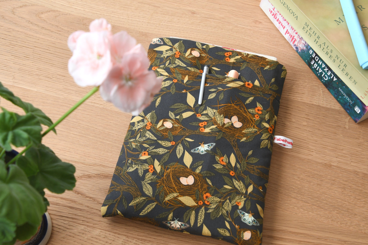 Handgefertigte, gepolsterte Buchtasche der Fabelhaften Werkstatt aus braunem Stoff mit einem detailreichen Natur-Motiv aus kleinen Vogelnestern und hellen Vogeleiern. Gut sichtbar ist das aufgesetzte Vorderfach für Zubehör auf der flach liegenden Hülle.